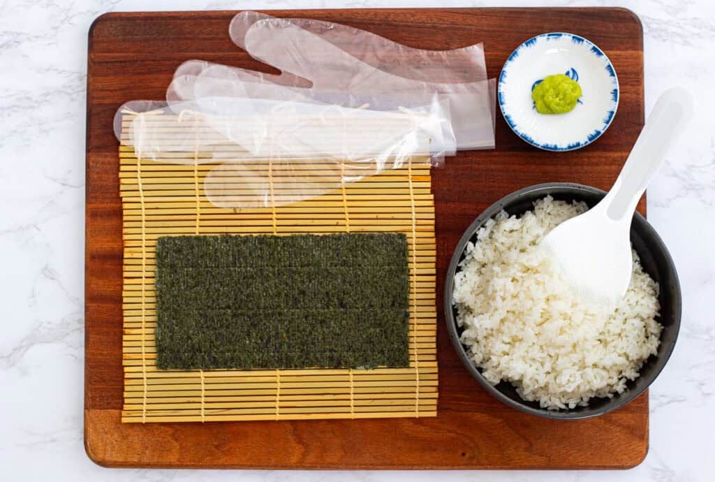 Easy maki roll recipe setup with bamboo mat, nori sheet, seasoned sushi rice, rice paddle, wasabi, and gloves ready for rolling hosomaki