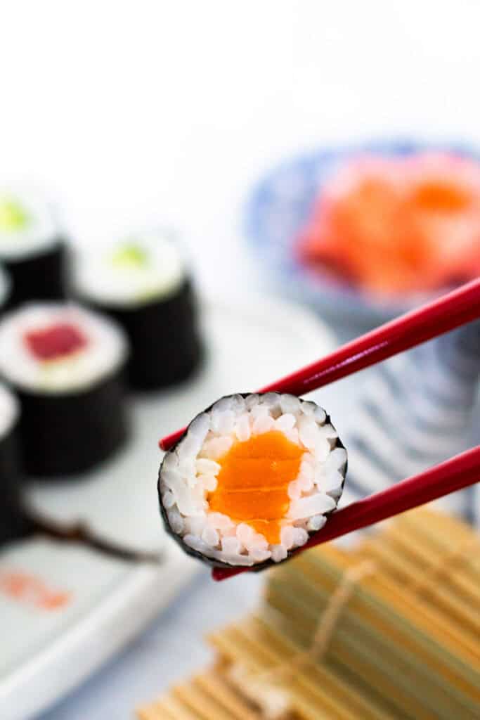 Close-up of salmon maki roll held with red chopsticks, with more sushi and pickled ginger in the background
