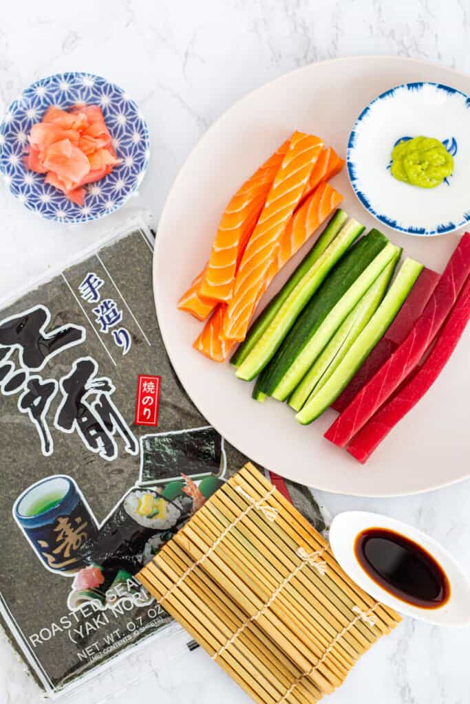 Ingredients for maki rolls, including salmon, tuna, cucumber, nori sheets, pickled ginger, wasabi, and soy sauce with bamboo mat