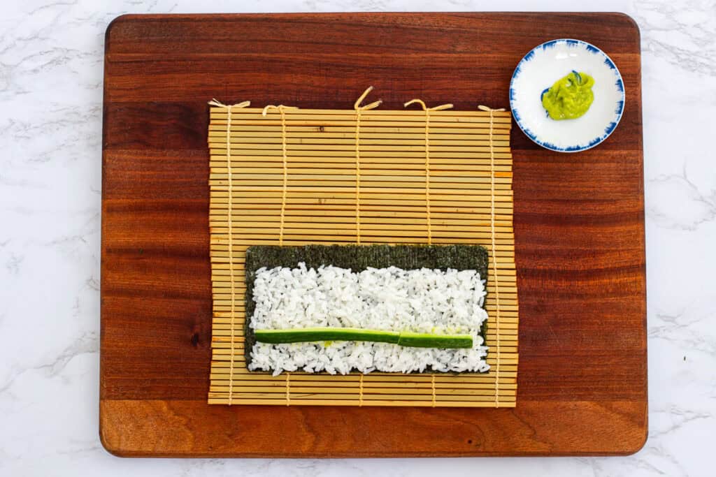Sushi rice evenly spread on nori sheet with wasabi paste and cucumber strip, ready to roll hosomaki
