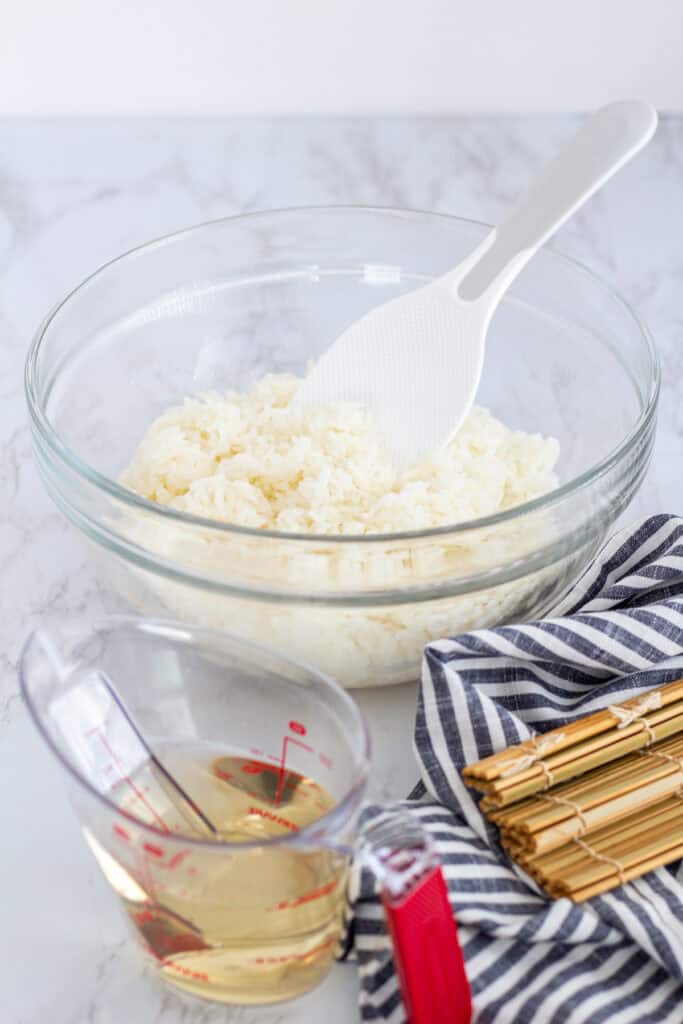 Seasoning sushi rice in a glass bowl with rice paddle, measuring cup of vinegar mixture, and bamboo mats on the side