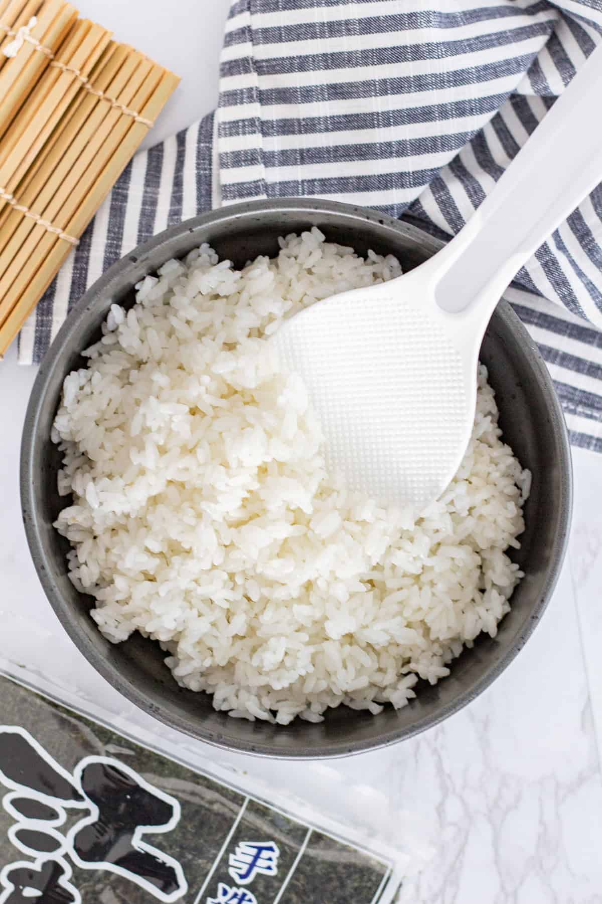 Seasoned sushi rice in a bowl with a rice paddle, ready to roll with nori sheets and bamboo mat nearby