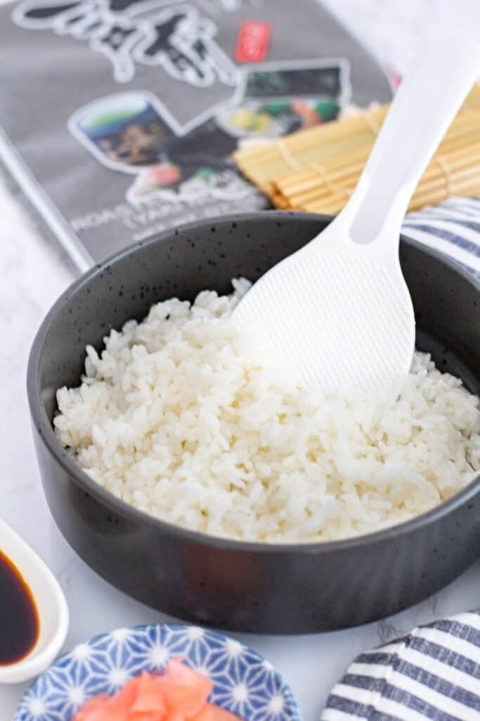 Bowl of seasoned sushi rice with rice paddle, nori sheets, bamboo mat, soy sauce, and pickled ginger ready for rolling sushi