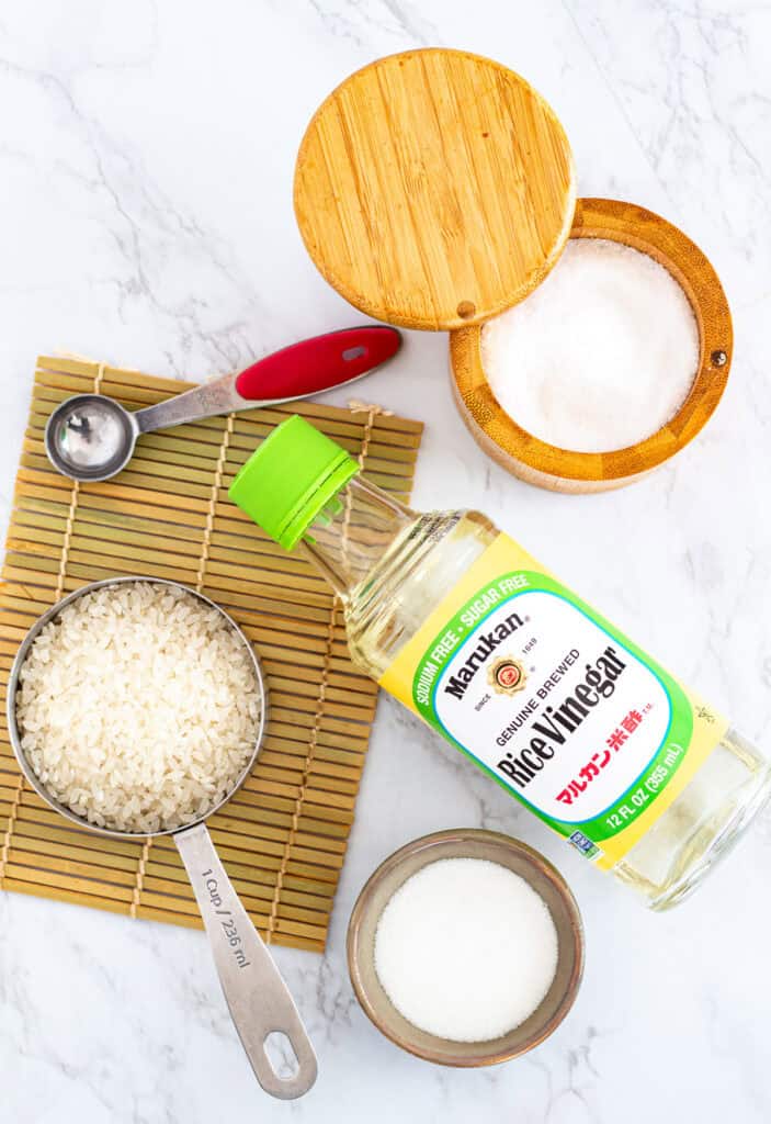 Ingredients for sushi rice seasoning, including rice vinegar, sugar, salt, and short-grain rice, displayed on a bamboo mat