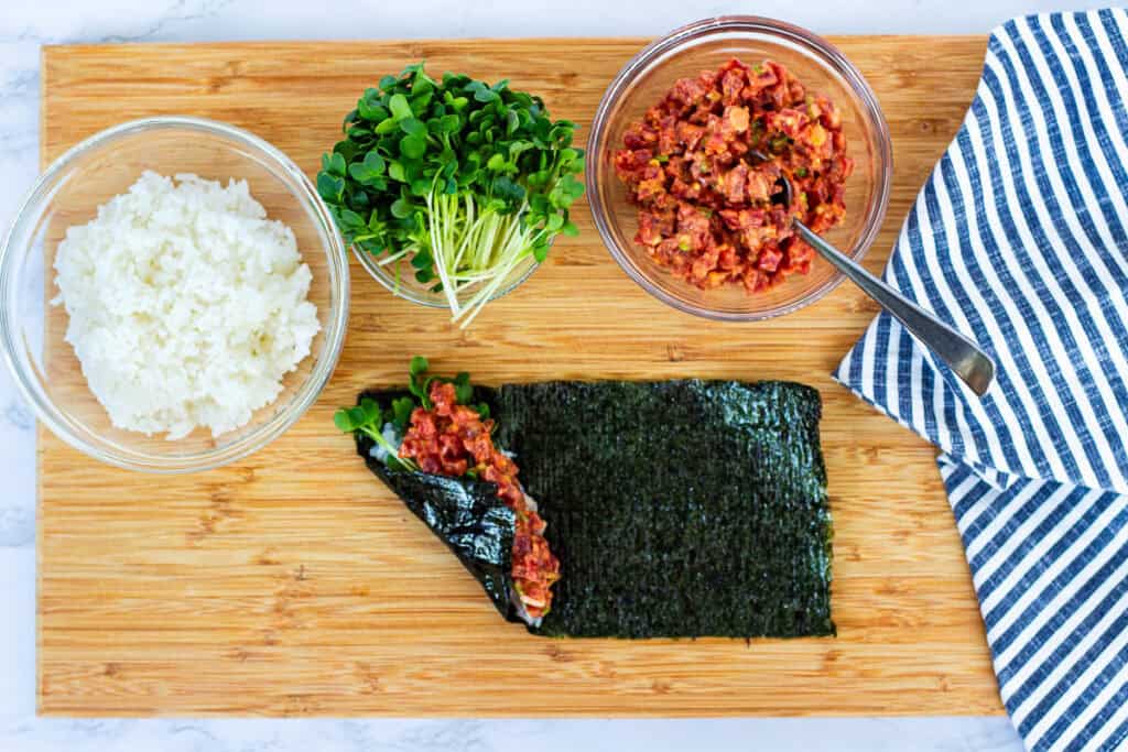 Spicy tuna hand roll being assembled with rice, sprouts, and spicy tuna filling, partially rolled on nori sheet