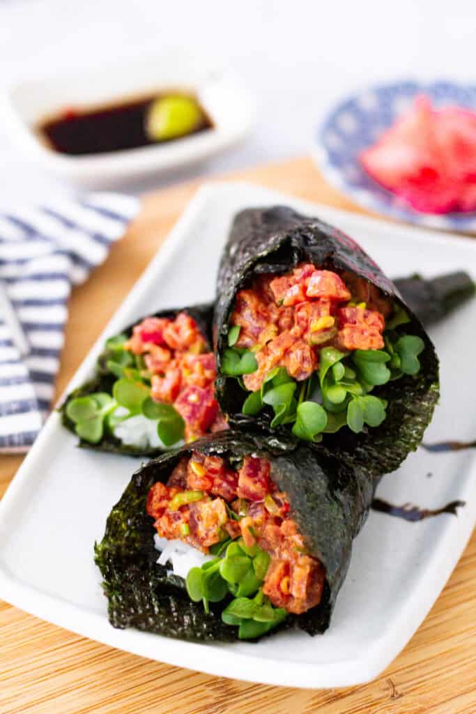 Three spicy tuna temaki hand rolls plated with sushi rice, spicy tuna, radish sprouts, and crisp nori, served with soy sauce and pickled ginger