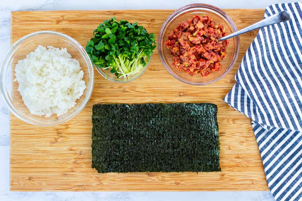 Ingredients arranged for making spicy tuna hand roll recipe, with seasoned sushi rice, spicy tuna filling, radish sprouts, and nori sheet on a board