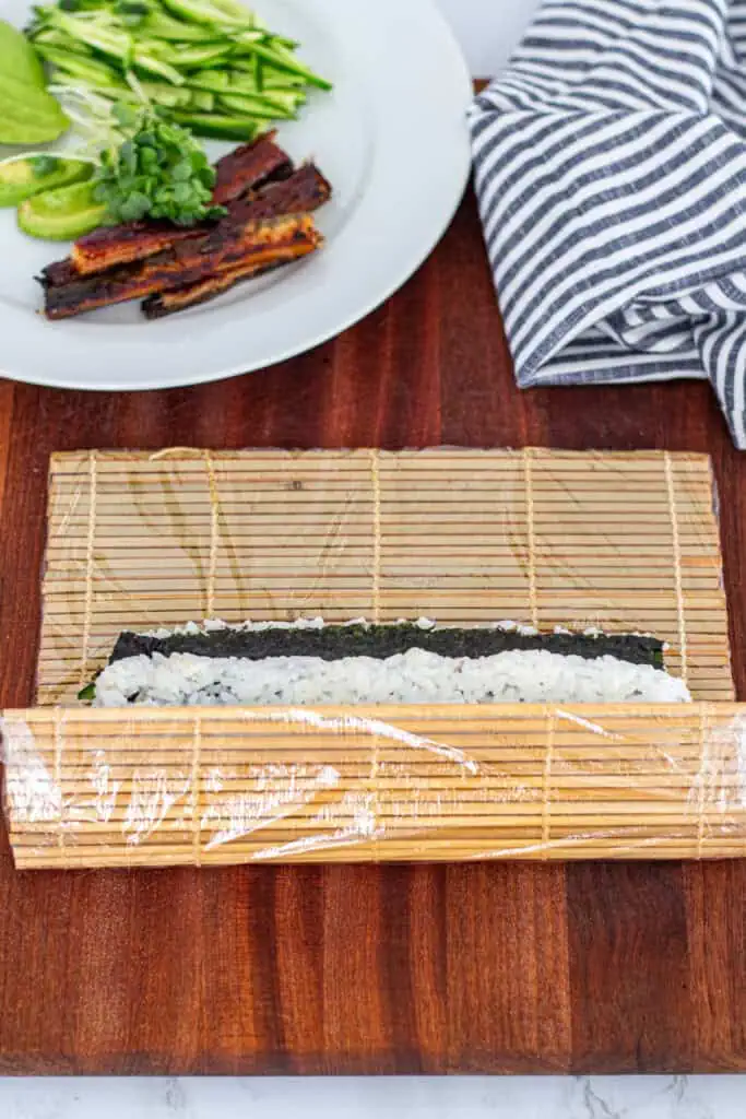 Unagi uramaki being rolled tightly with a bamboo sushi mat wrapped in plastic wrap