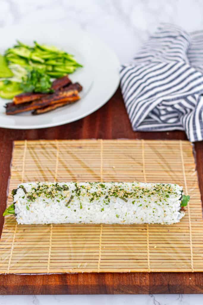 Unagi uramaki roll fully shaped and topped with furikake, resting on a bamboo mat before slicing