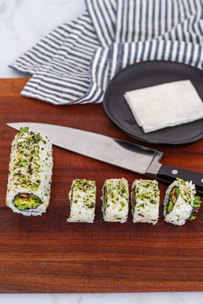 Unagi uramaki roll partially sliced on a wooden cutting board, with a sharp knife and damp towel for clean cuts