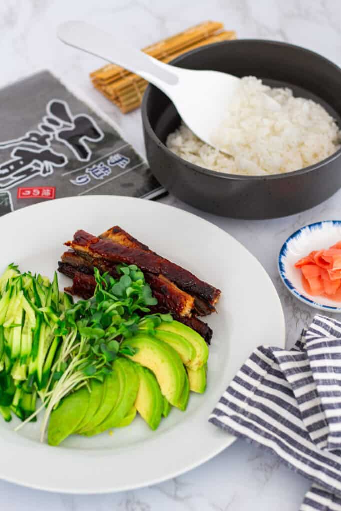 A plate of sliced cucumber, avocado, radish sprouts, and baked unagi with a bowl of seasoned rice and pickled ginger, ready for making uramaki rolls