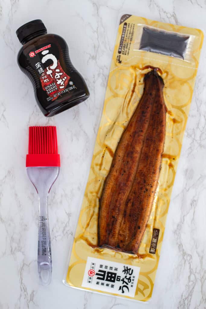 Packaged grilled unagi, bottle of Kikkoman unagi sauce, and silicone brush on marble surface, ready to make unagi uramaki rolls