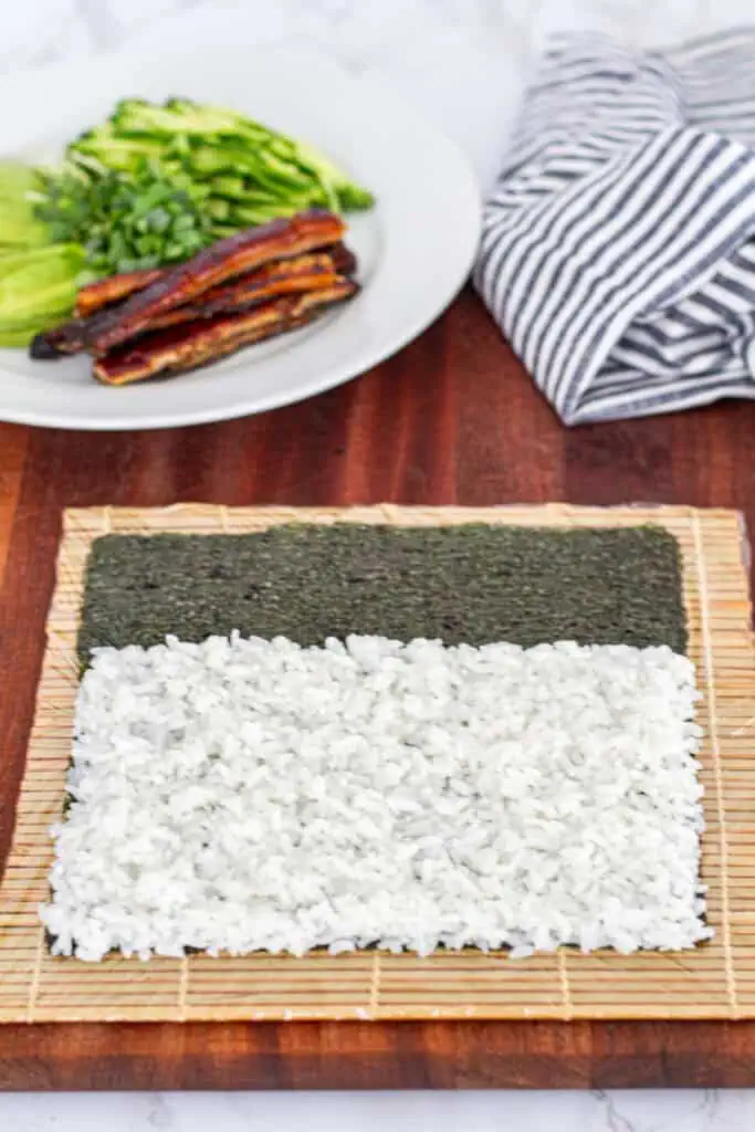 A bamboo mat with a sheet of nori partially covered in sushi rice, ready to be flipped and filled for an unagi uramaki roll