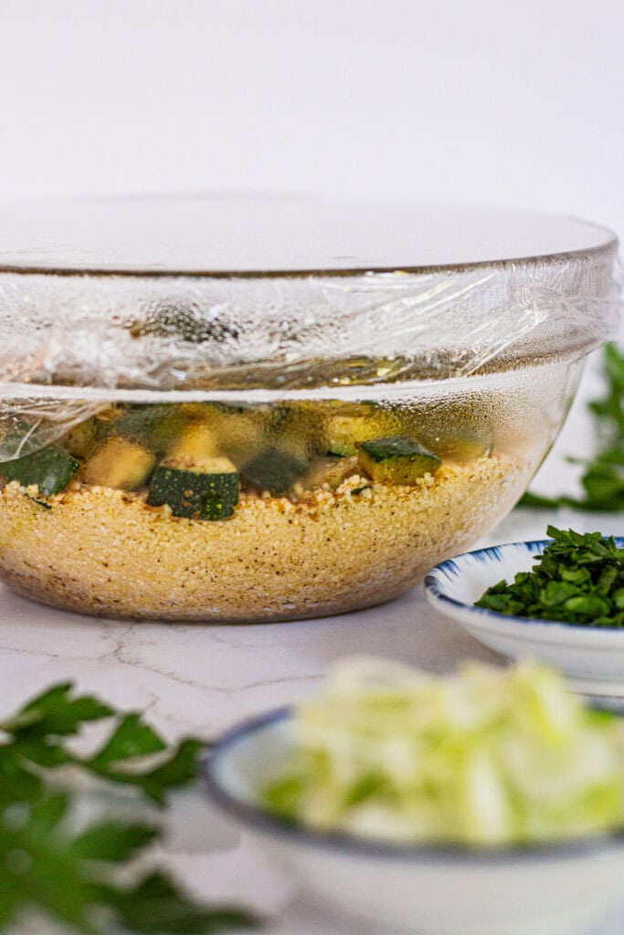 Glass bowl of couscous and sautéed zucchini covered with plastic wrap to steam with garam masala