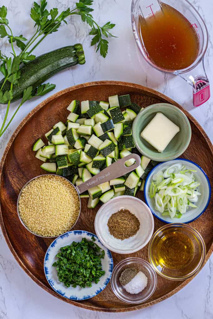 Ingredients for zucchini couscous with garam masala arranged on a wooden tray, including diced zucchini, couscous, parsley, scallions, broth, butter, oil, and spices.