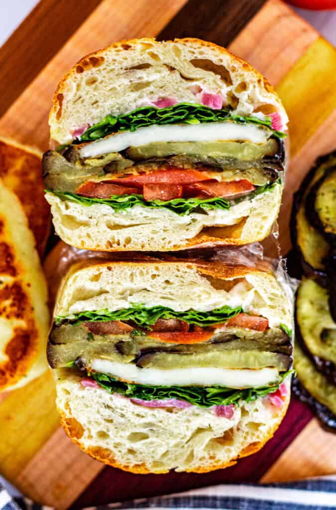 Overhead view of roasted eggplant sandwich cut in half showing layers of halloumi, tomato, greens, and garlic spread