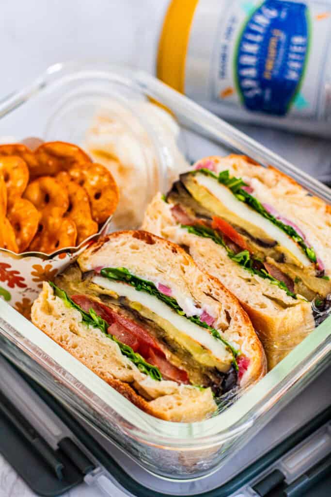 Mediterranean eggplant and halloumi sandwich packed in a glass lunchbox with pretzel crisps and garlic dip