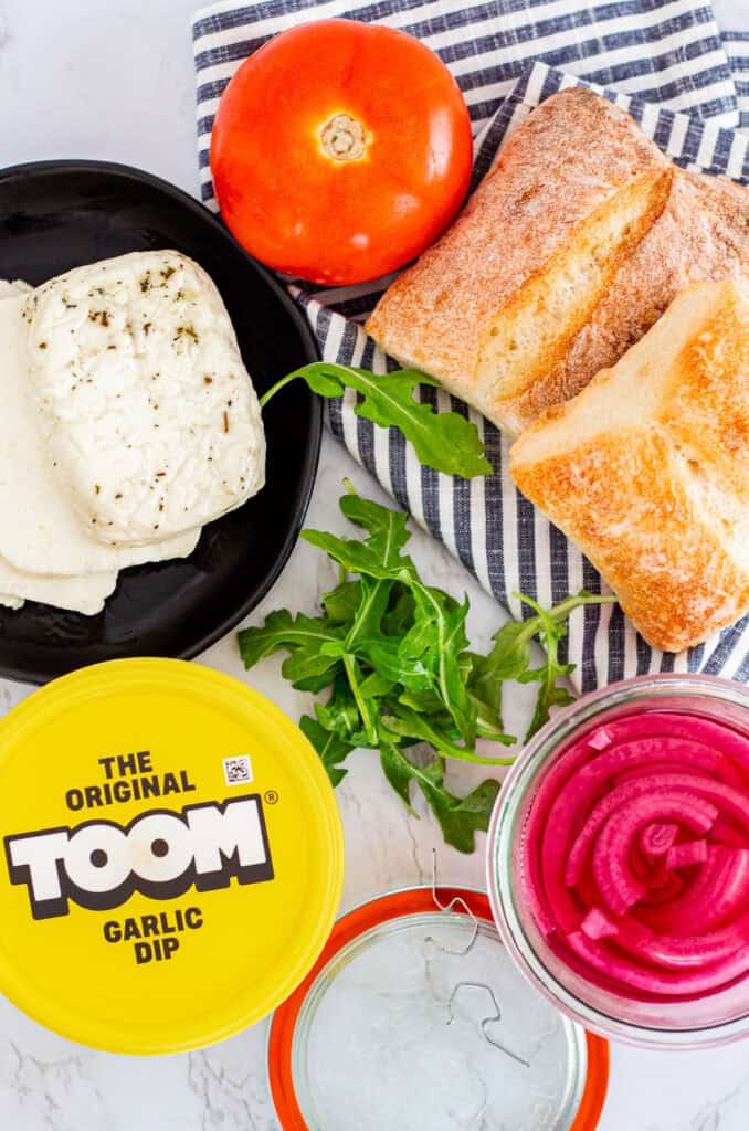 Ingredients for Mediterranean eggplant sandwich including halloumi, tomato, ciabatta rolls, garlic dip, arugula, and pickled onions