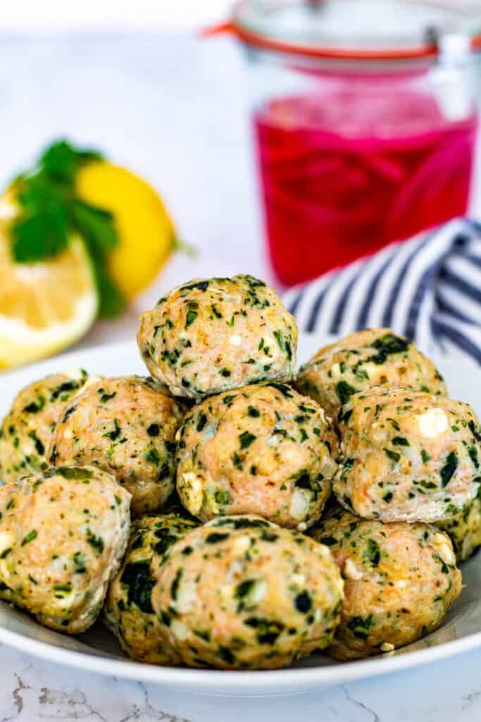 Oven baked Greek chicken meatballs with feta and herbs served in a white bowl with lemon and pickled onions in the background