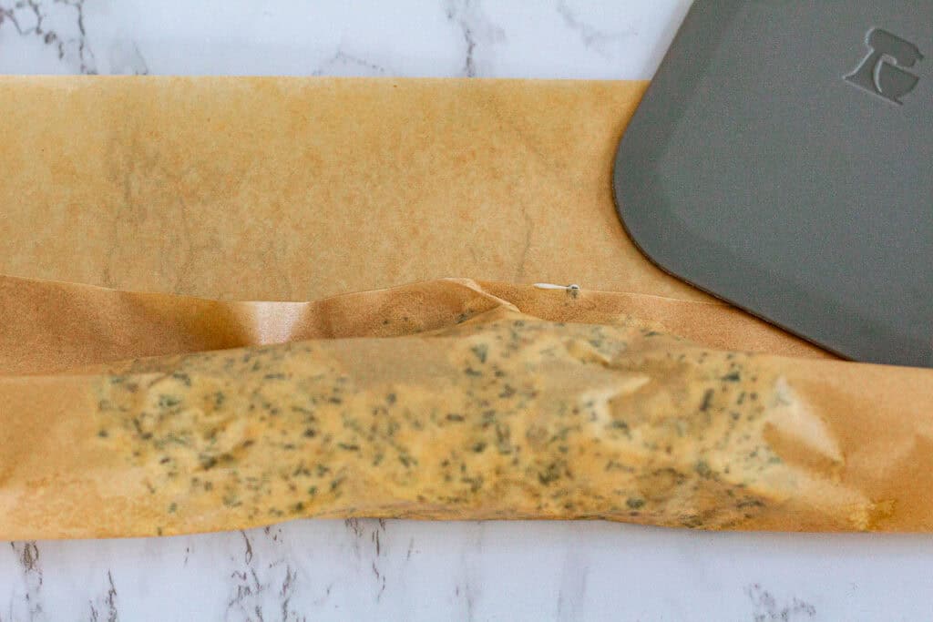 Rolling and wrapping homemade herb butter in parchment paper before refrigerating or freezing.