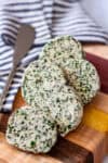 Sliced herb butter made with fresh rosemary, sage, thyme, and parsley, arranged on a wooden cutting board with a butter knife and striped cloth in the background.