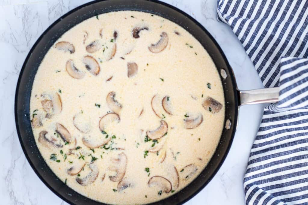 Cream, broth, wine, and mushrooms simmering in a skillet with herbs for marsala cream sauce.