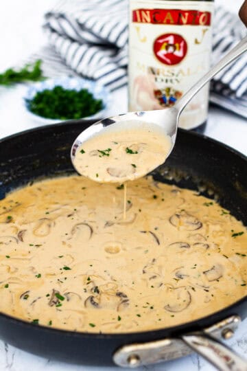 Spoon lifting marsala cream sauce with mushrooms from a skillet, showing creamy texture.