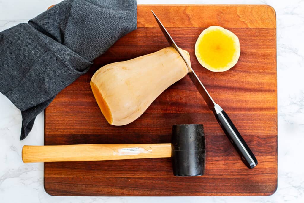 Butternut squash on a cutting board with the top removed, a knife halfway through the squash, and a mallet for safe cutting.