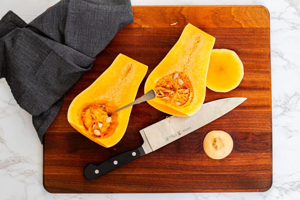Two butternut squash halves on a cutting board with seeds being scooped out using a spoon.