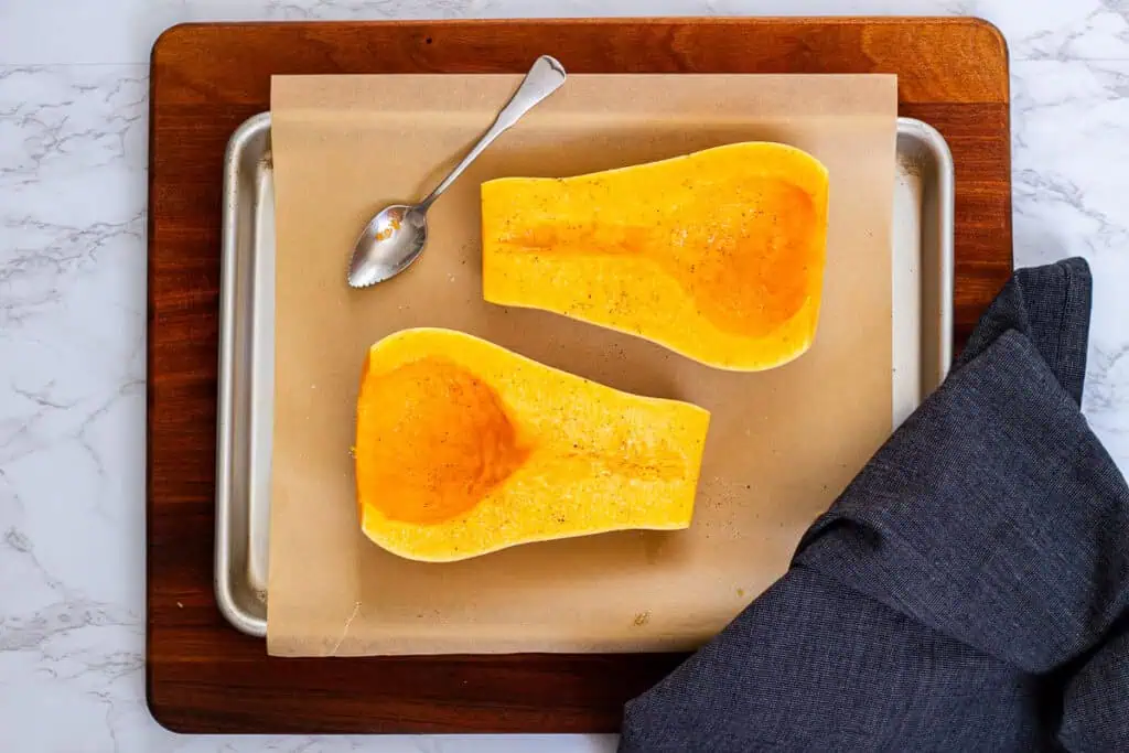 Two butternut squash halves on a parchment-lined baking sheet, seasoned with oil, salt, and pepper.