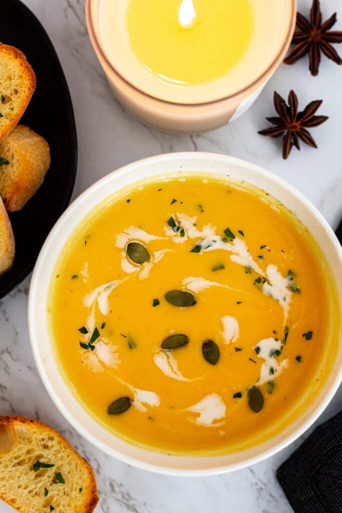 Close-up of creamy butternut squash bisque garnished with pumpkin seeds, fresh herbs, and a swirl of cream, served with crusty bread, with a lit candle and star anise in the background.