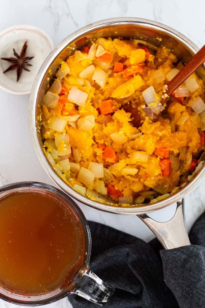 Pot filled with roasted butternut squash, carrots, and onions, ready to be cooked for creamy butternut squash bisque, with a small bowl of star anise and a measuring cup of broth on the side.