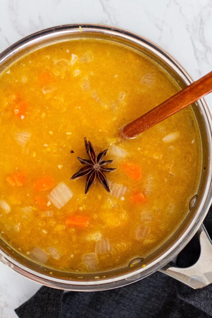 Pot of butternut squash bisque base simmering in broth with carrots, onions, and a star anise pod, stirred with a wooden spoon.