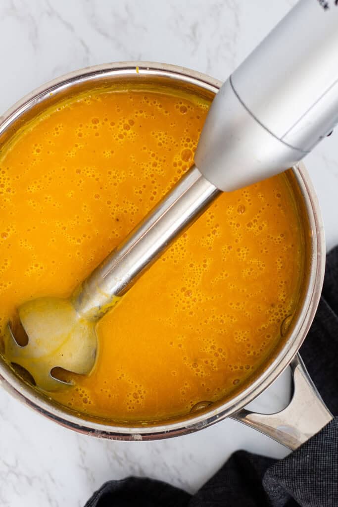 Immersion blender blending cooked butternut squash, carrots, and onions into a smooth bisque inside a stainless steel pot.