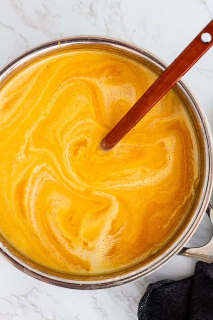 Wooden spoon stirring cream into pureed butternut squash bisque, creating a golden swirl pattern in the pot.