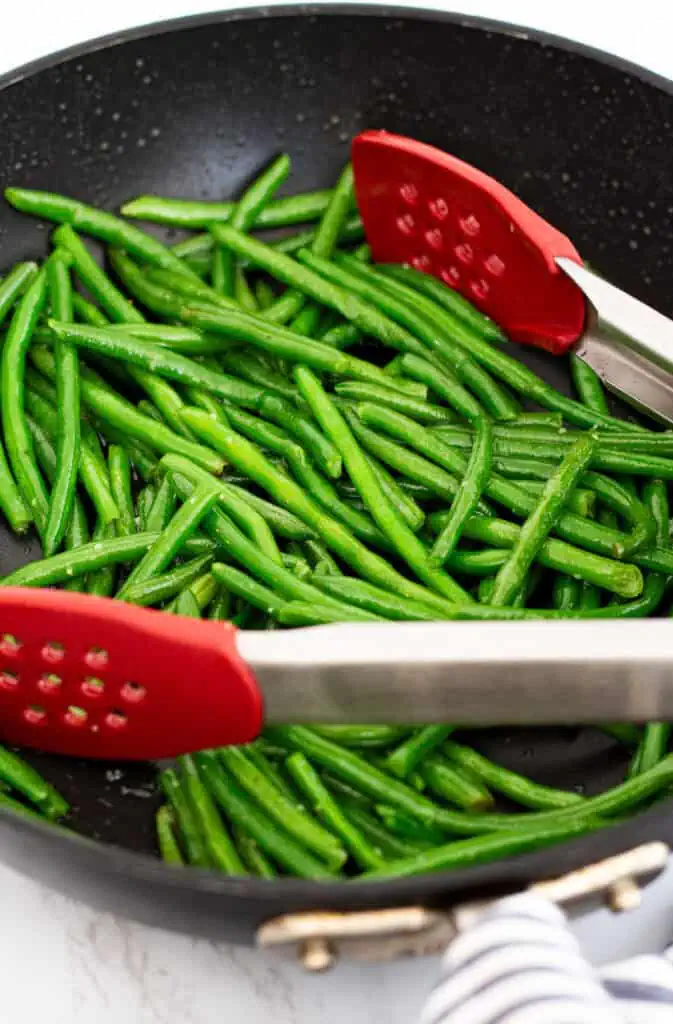 Green beans sautéing in a black skillet with olive oil and garlic salt, stirred with red tongs