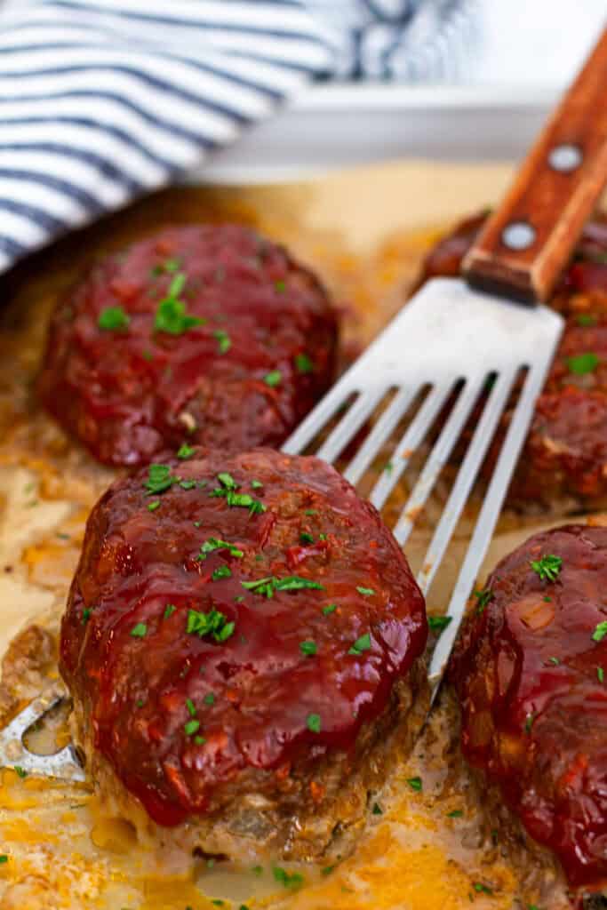 Close-up of mini meatloaf with shiny ketchup glaze, garnished with chopped parsley and lifted with a spatula