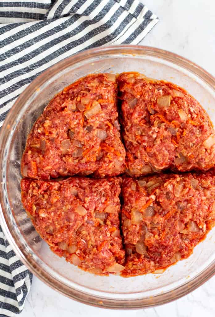 Raw meatloaf mixture with onions and carrots divided into four portions in a glass mixing bowl