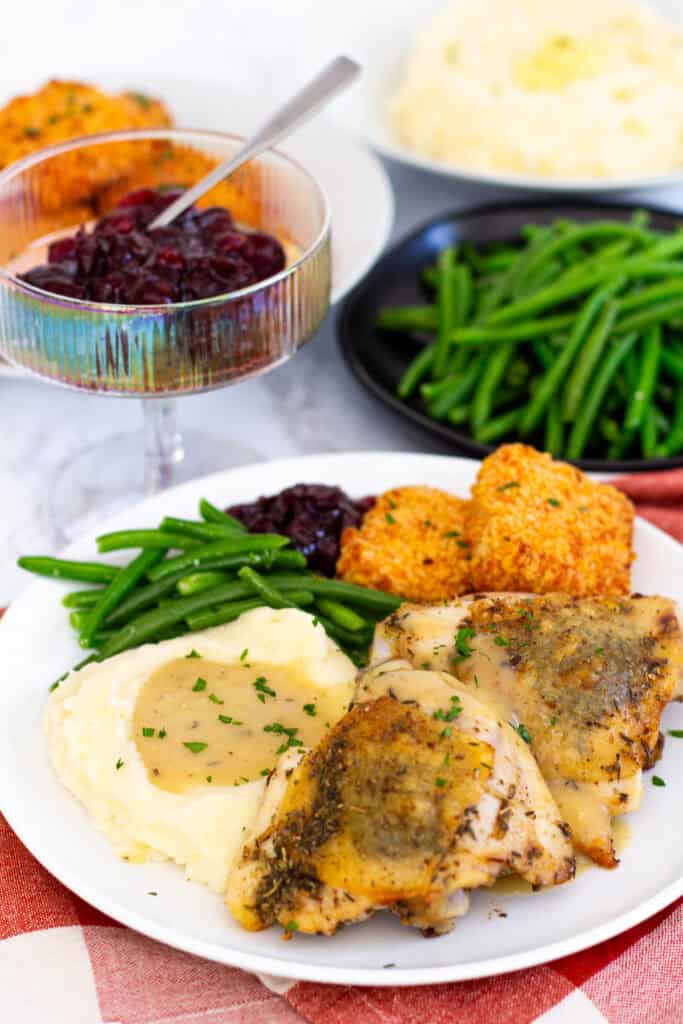 Plate with roasted chicken thighs, mashed potatoes and gravy, green beans, cranberry sauce, and mac and cheese bites for a Thanksgiving meal for two.