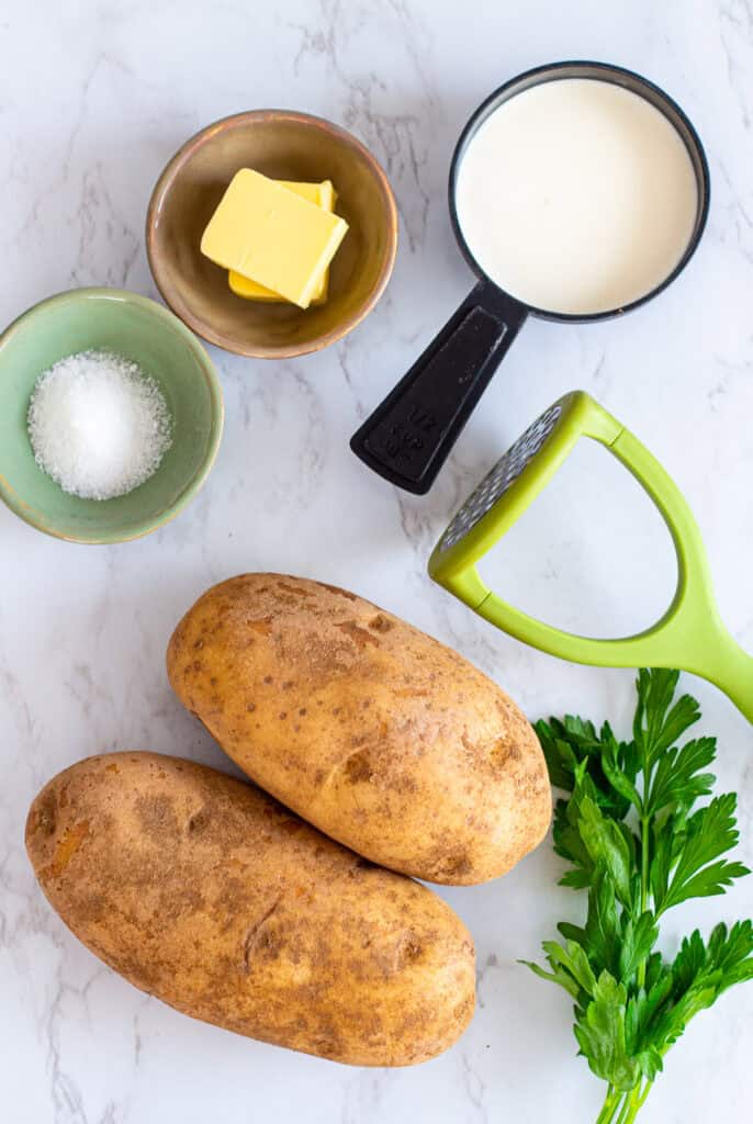 Russet potatoes, butter, cream, salt, and parsley on marble counter with a potato masher