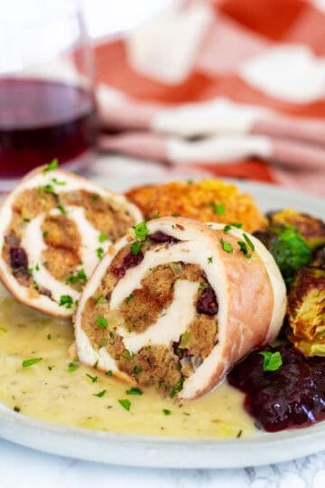 Sliced stuffed chicken breast with stuffing served on a plate with gravy, cranberry sauce, and roasted vegetables.