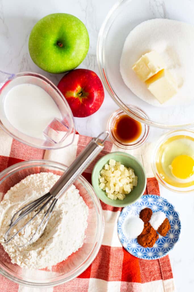 Ingredients for mini spiced apple cake including apples, butter, sugar, flour, egg, spices, and candied ginger arranged on red checkered cloth