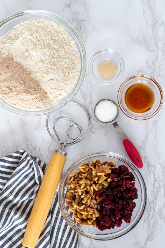 Flour, whole wheat flour, sourdough discard, honey, salt, instant yeast, walnuts, and dried cranberries arranged in bowls for cranberry walnut bread.