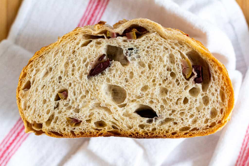 A sliced loaf of sourdough olive bread showing a soft, airy crumb with pieces of green and kalamata olives.