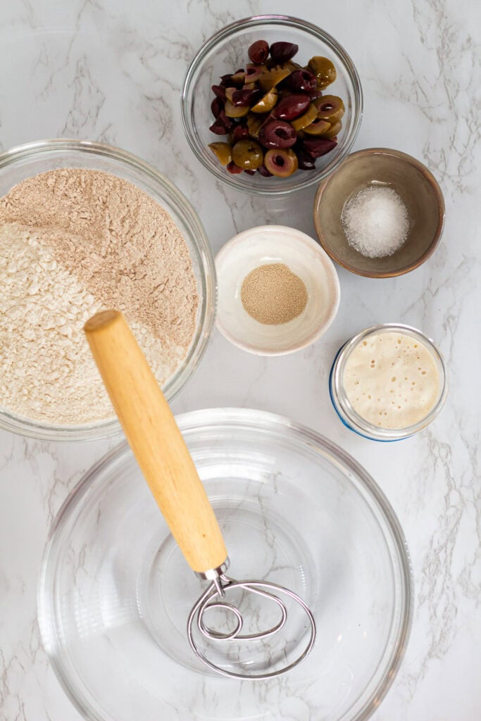 A top-down view of sourdough olive bread ingredients, including flour, olives, salt, yeast, and starter arranged around a glass bowl with a dough whisk.
