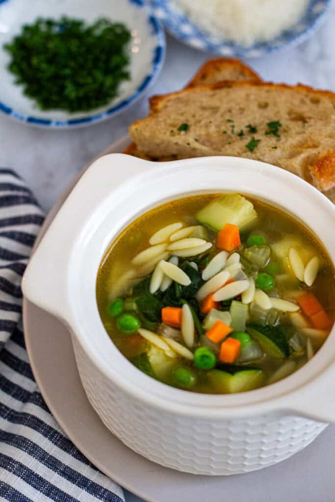A warm bowl of vegetable orzo soup with carrots, peas, zucchini, spinach, and tender orzo in broth, served with toasted bread.