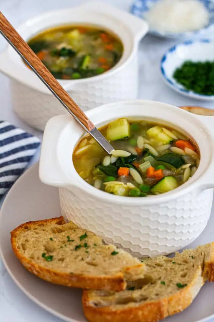 Two bowls of vegetable orzo soup on a table setting, with bread slices and fresh herbs in the background.