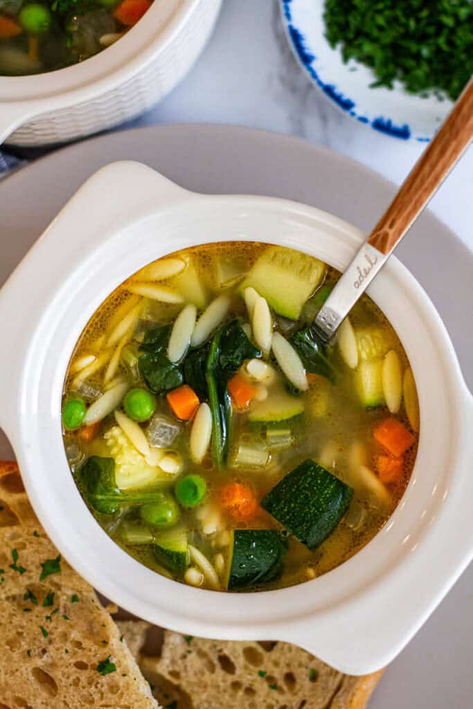 Close up view of vegetable orzo soup showing tender orzo, zucchini, carrots, peas, and spinach in broth.