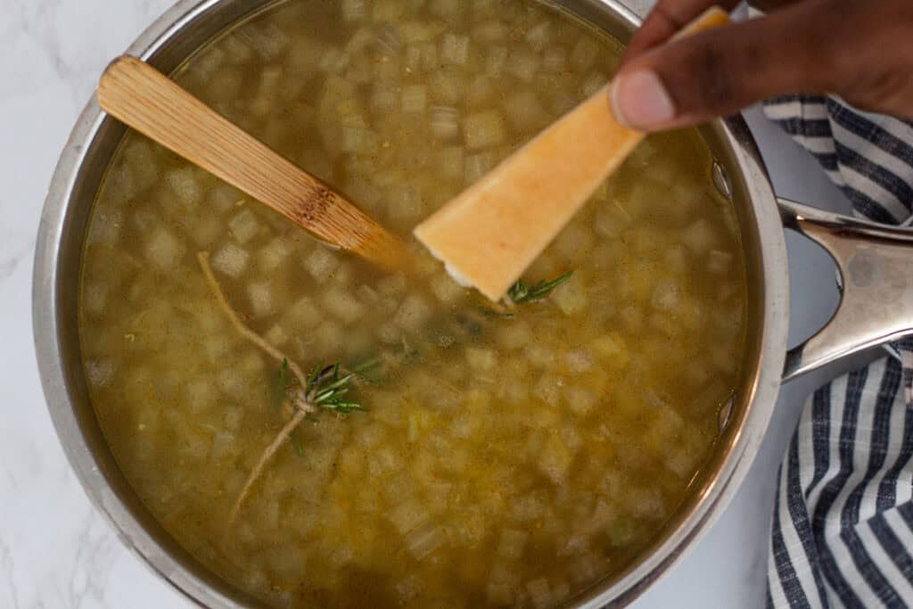 A parmesan rind being added to simmering broth with rosemary.
