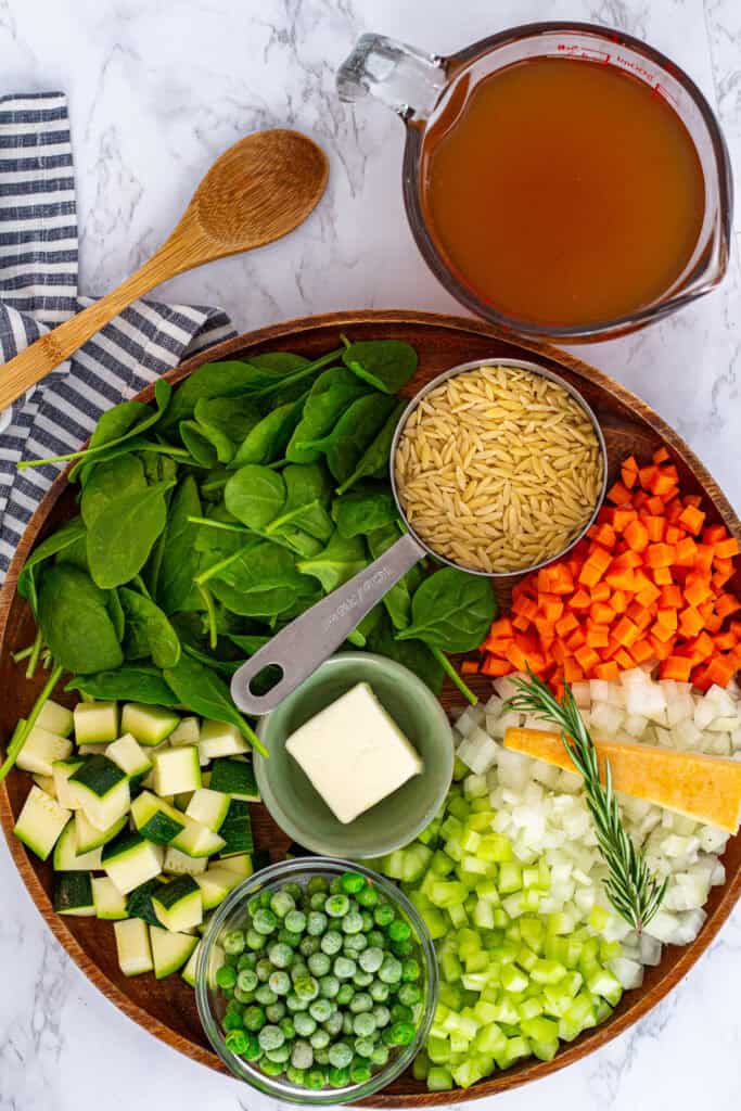 Vegetable orzo soup ingredients arranged on a wooden board, including vegetables, broth, and orzo.